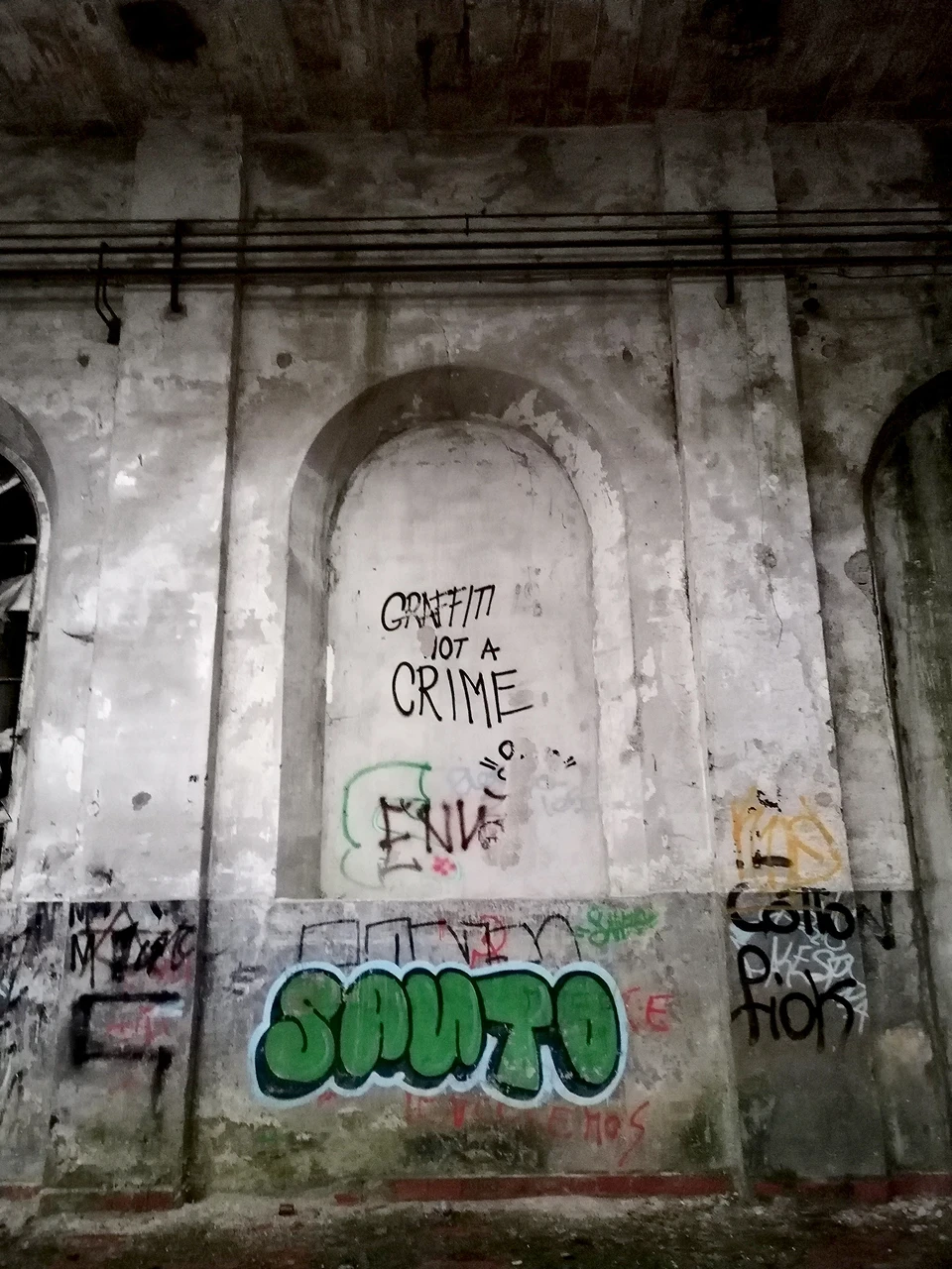 Interno di una struttura industriale abbandonata con archi strutturali e diversi graffiti, tra cui la scritta "Graffiti is not a crime".