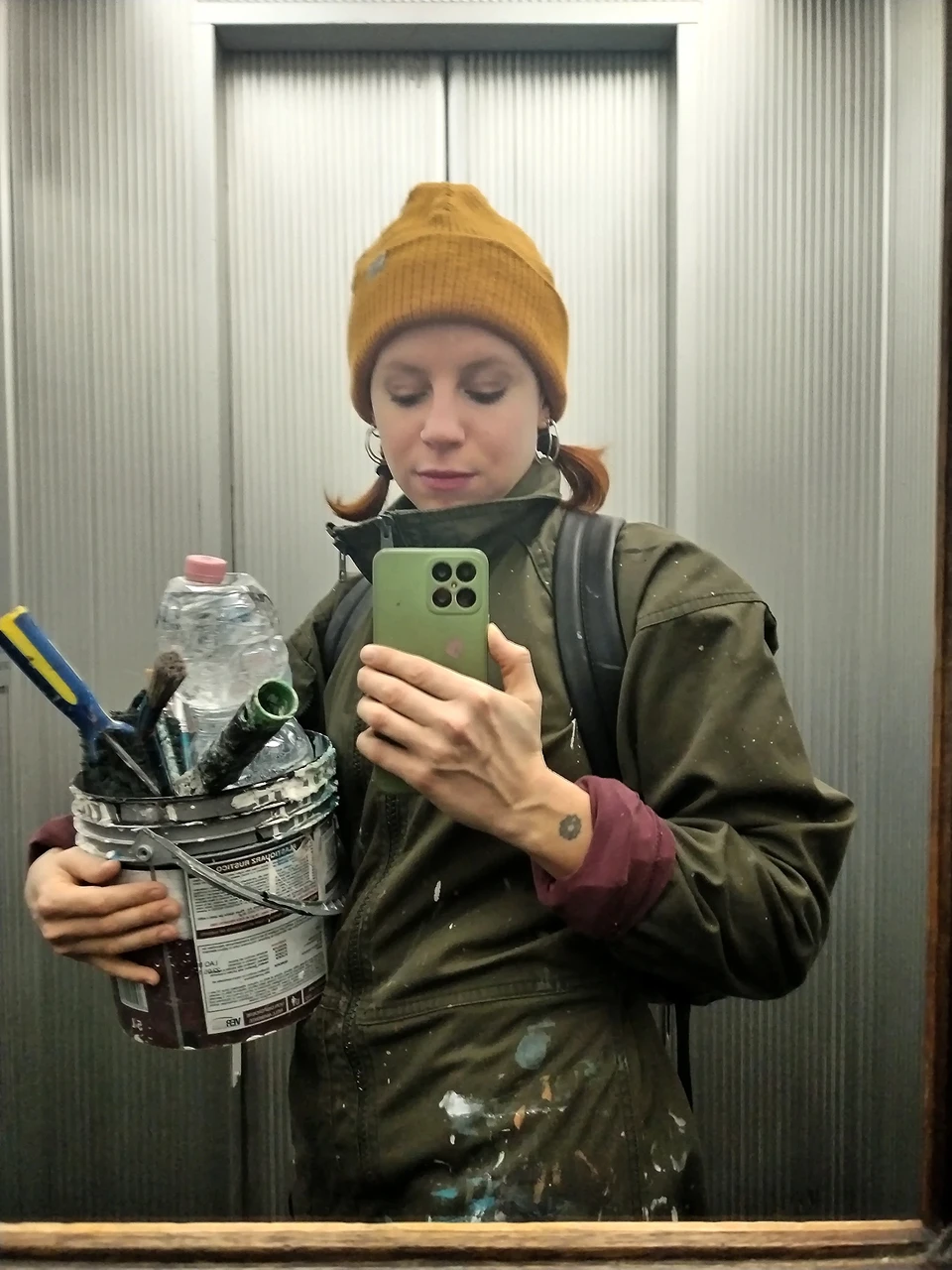 Selfie di Rosmunda in un ascensore industriale, con indosso un berretto giallo e un secchio di vernice pieno di pennelli e attrezzi da lavoro.